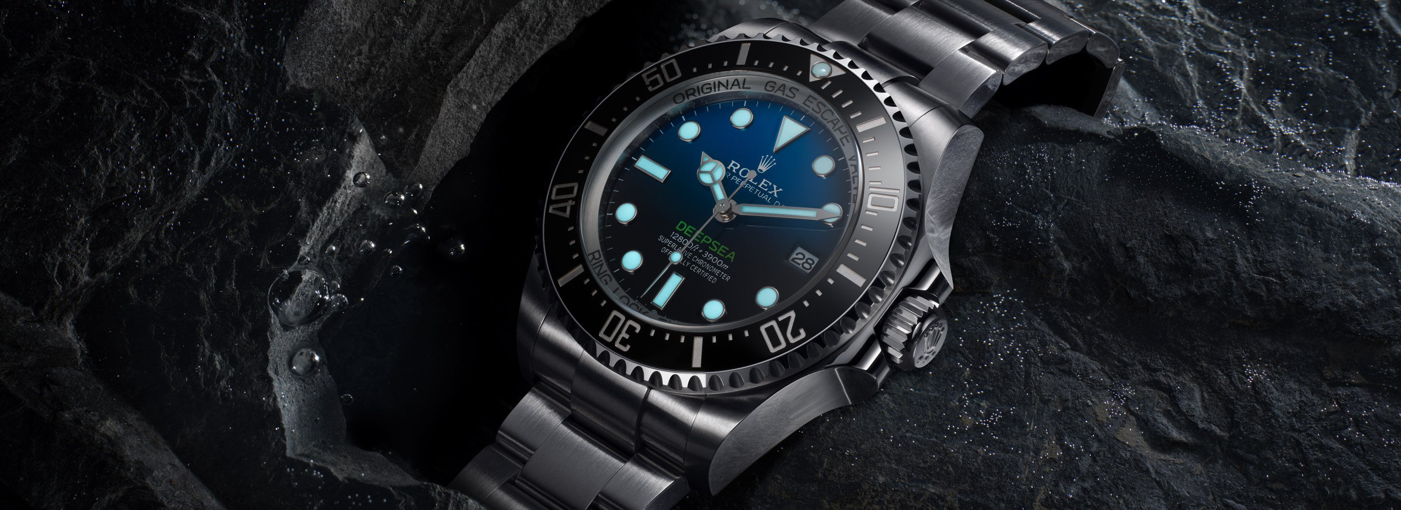 Rolex Deepsea in Oystersteel with D-blue dial and black Cerachrom bezel on an Oyster bracelet, resting on dark wet rocks with water droplets and Chromalight luminescence glowing from the dial