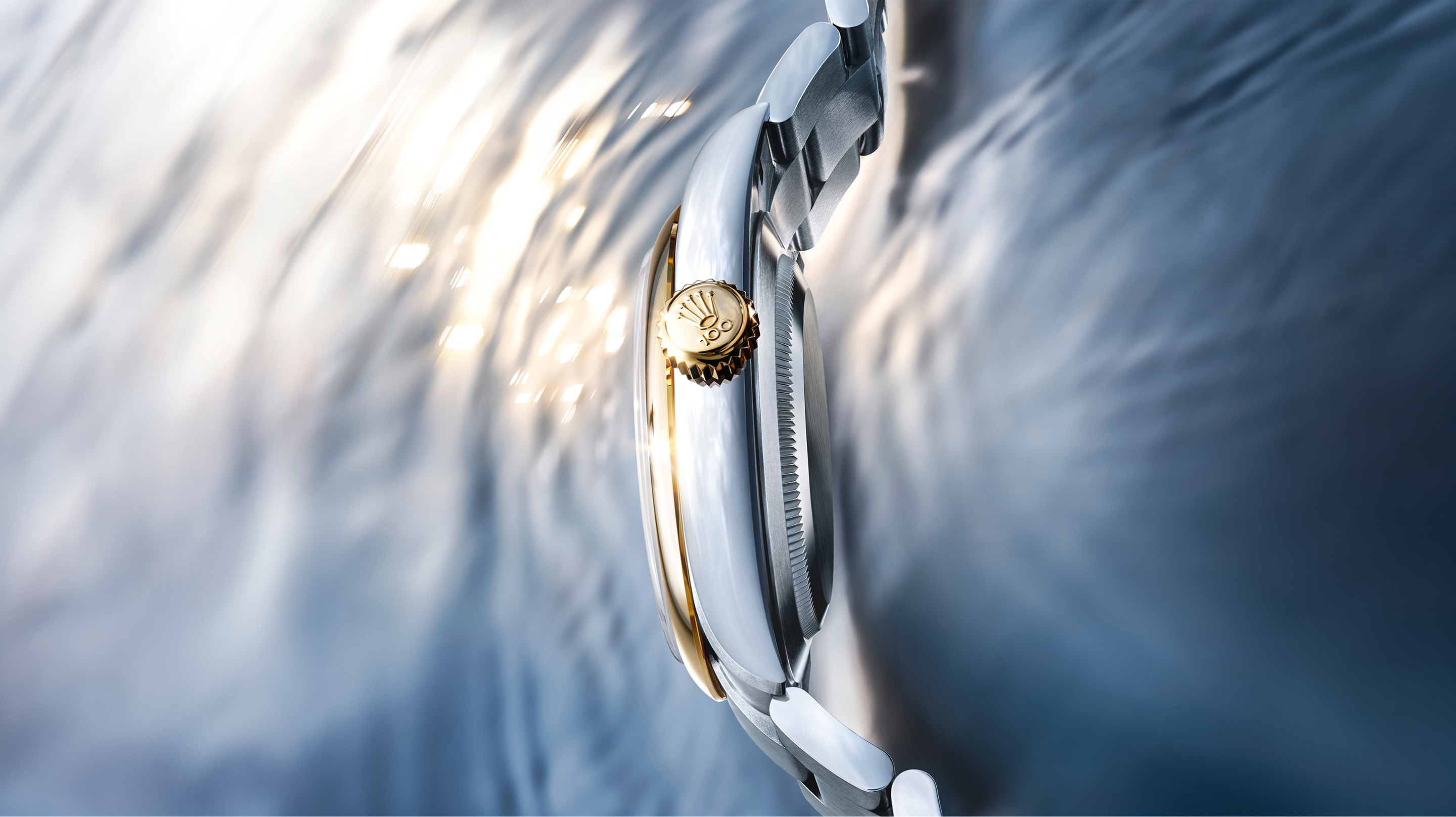 Side profile of the Rolex Oyster Perpetual 100th anniversary yellow Rolesor edition, showing the Twinlock winding crown with Rolex crown emblem against a moving water surface