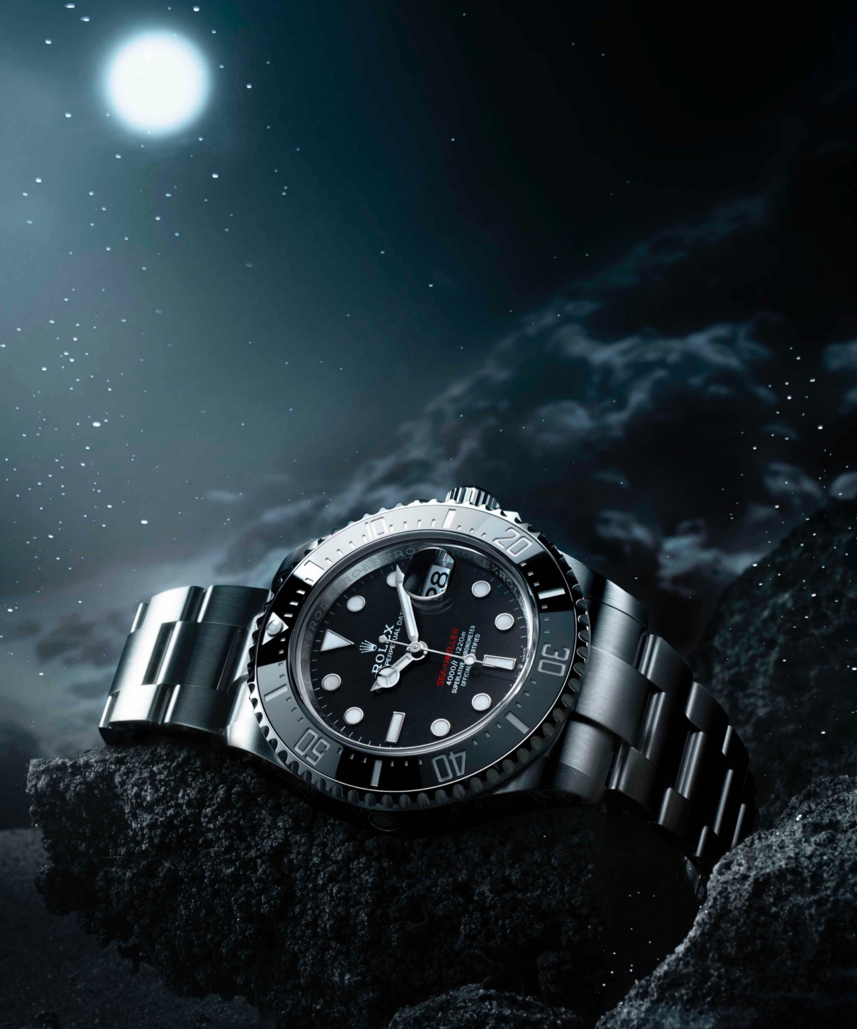 Rolex Sea-Dweller in Oystersteel with black dial and black Cerachrom bezel resting on dark coastal rocks beneath a starry night sky with a full moon