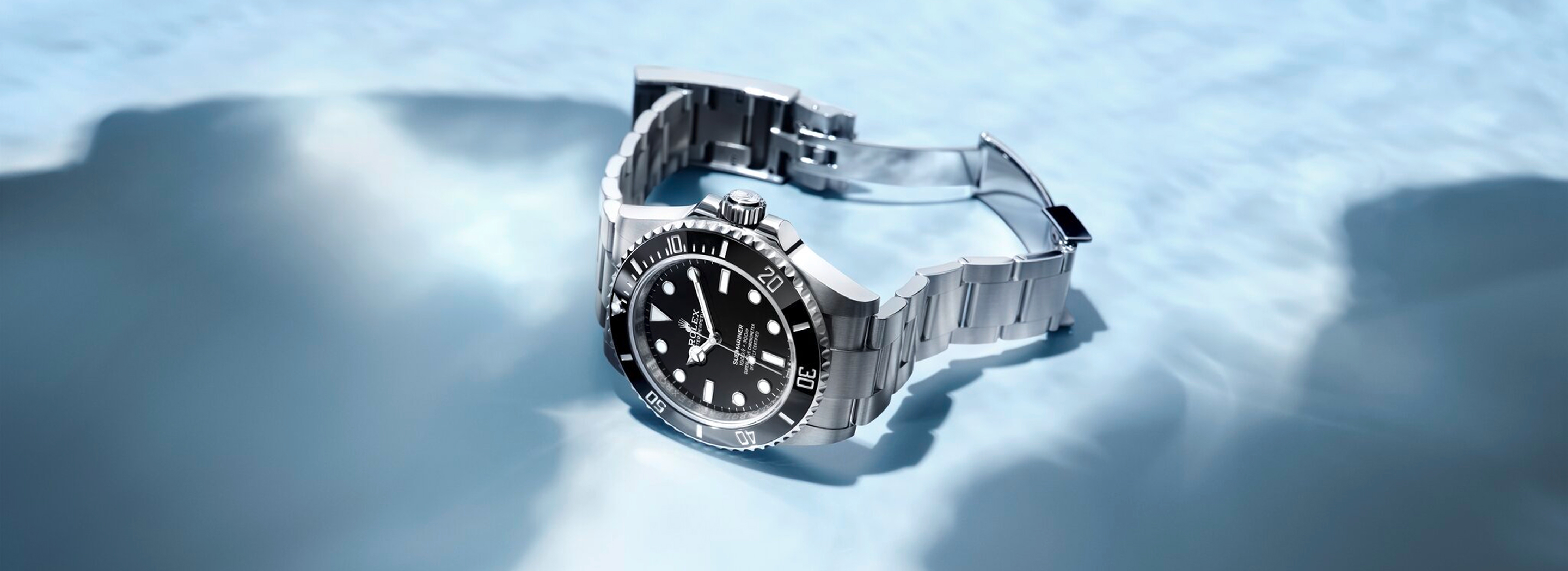 Rolex Submariner in Oystersteel with black dial and black Cerachrom bezel on an Oyster bracelet with Oysterlock clasp open, resting on an icy surface against a blue background