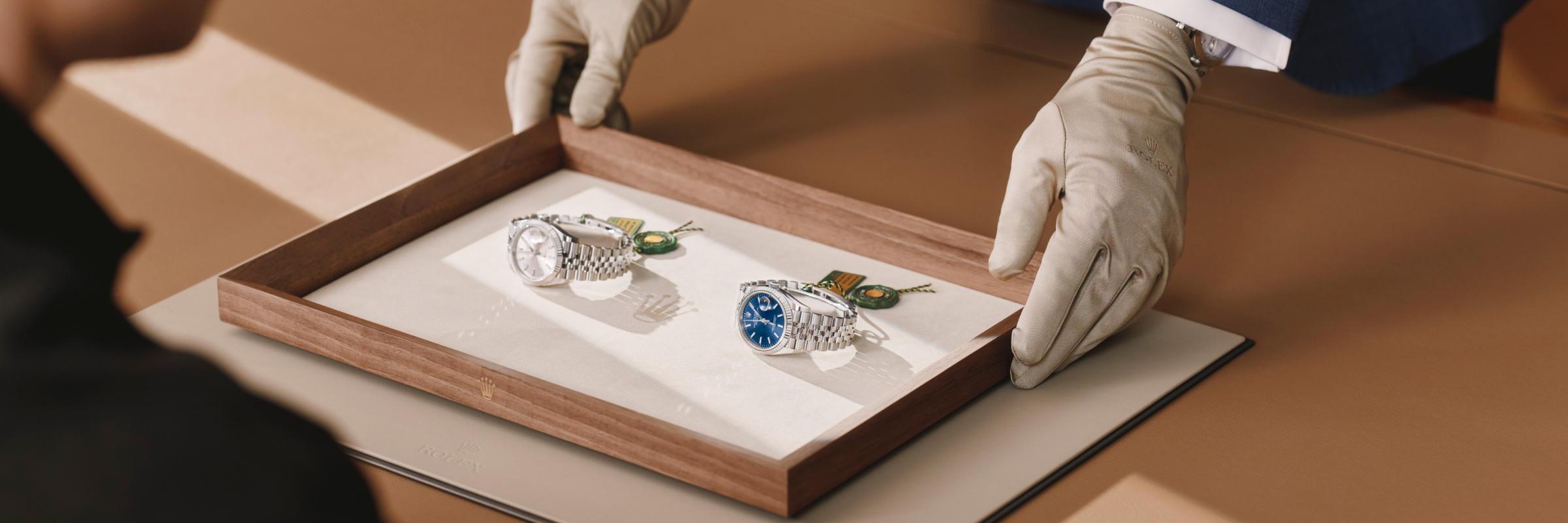 Gloved hands present two luxury wristwatches—one with a white dial and one with a blue dial—on a wooden display tray in a boutique setting.
